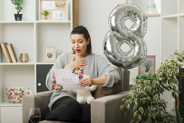 Pleased putting hand on heart beautiful girl on happy women day holding and reading greeting card sitting on armchair in living room