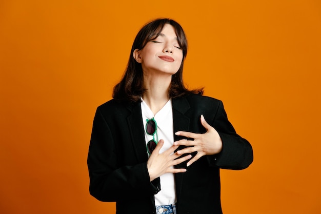 Pleased putting hands on heart young beautiful female wearing black jacket isolated on orange background