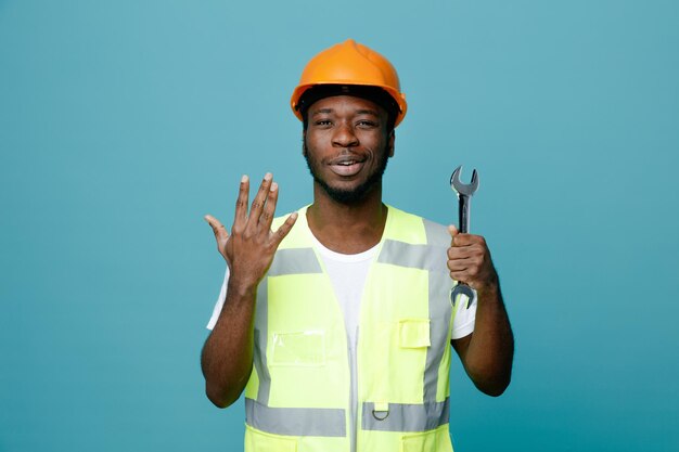 Pleased raised hand young african american builder in uniform holding open end wrench isolated on blue background