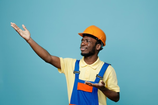 Pleased raised hand young african american builder in uniform isolated on blue background