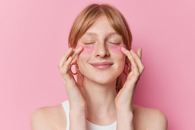 Pleased redhaired female model undergoes beauty procedures appies hydrogel patches under eyes to remove puffiness and fine lines wears casual t shirt isolated over pink background Skin care