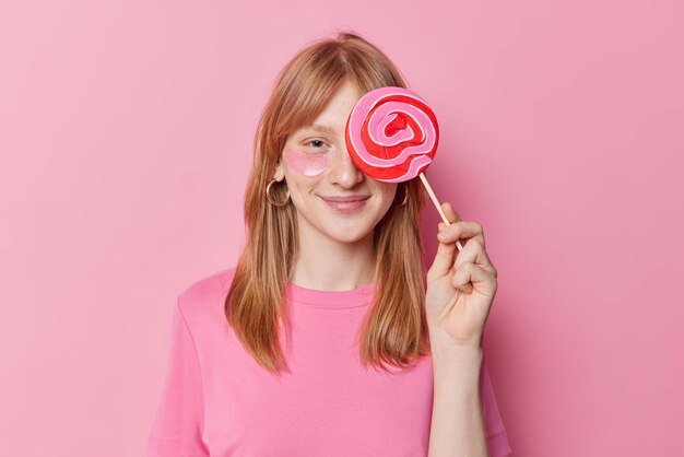 기뻐하는 빨간 머리 소녀는 크고 맛있는 사탕으로 눈을 가리고 미용 패치를 적용하고 분홍색 배경에 캐주얼 티셔츠 포즈를 취합니다.