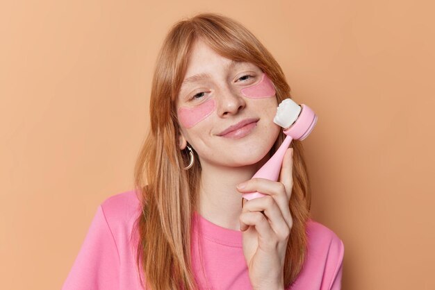 Pleased redhead girl uses face massager for skin care applies hydrogel patches under eyes to remove puffiness tilts head wears casual t shirt isolated over brown backgound Facial treatments