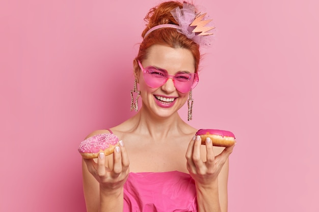 만족스러운 빨간 머리 여자는 긍정적 인 감정을 표현하고 행복하게 좋은 분위기에있는 두 개의 식욕을 돋우는 도넛 미소를 가지고 있습니다.