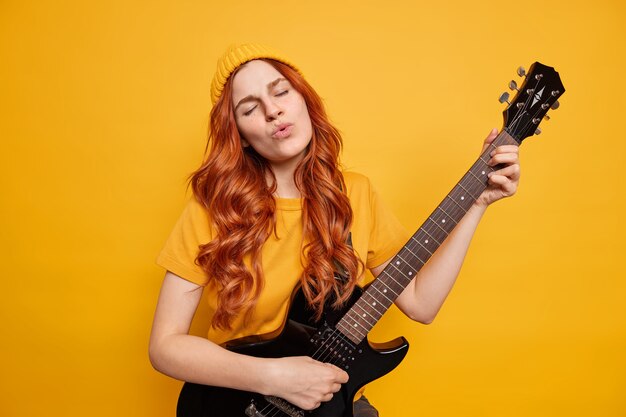 Pleased relaxed female guitarist plays bass guitar closes eyes from satisfaction sings song has red hair dressed in casual clothes prepares for musical tour 