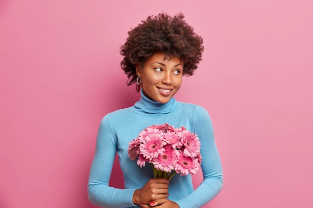 Pleased romantic African American woman holds bouquet of gerberas gets flowers