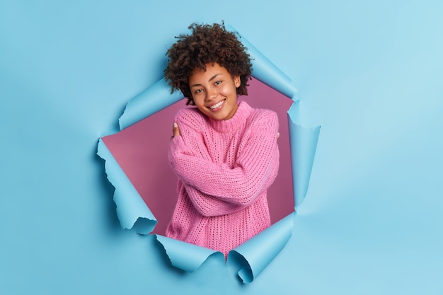 Pleased romantic happy young Afro American woman embraces herself needs feel warmth and love recalls lovely memory wears knitted sweater breaks through paper wall