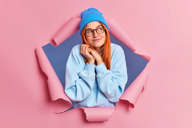 Pleased romantic redhead woman looks with dreamy expression aside keeps hands near face looks gladfully aside wears blue hat casual sweater.