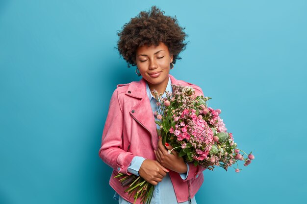 Pleased romantic woman gets beautiful bouquet as present, closes eyes and smiles gently, dressed in pink jacket