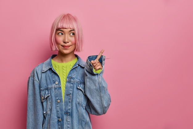 Free photo pleased rouge asian girl with bob hair, dressed in oversized denim jacket, points on copy space, advertises place,