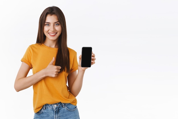 Pleased satisfied attractive stylish girl in yellow tshirt showing mobile phone display smartphone application make thumbup and smiling in approval agreement give recommendation