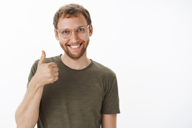 Pleased and satisfied excited funny european male with bristle in glasses and green casual t-shirt showing thumbs up and smiling joyfully approving good idea