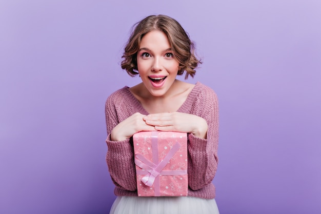 Donna dai capelli corti piacevole in posa con scatola regalo rosa carino e sorridente. incantevole ragazza riccia che gode del servizio fotografico con regalo di capodanno sul muro viola.