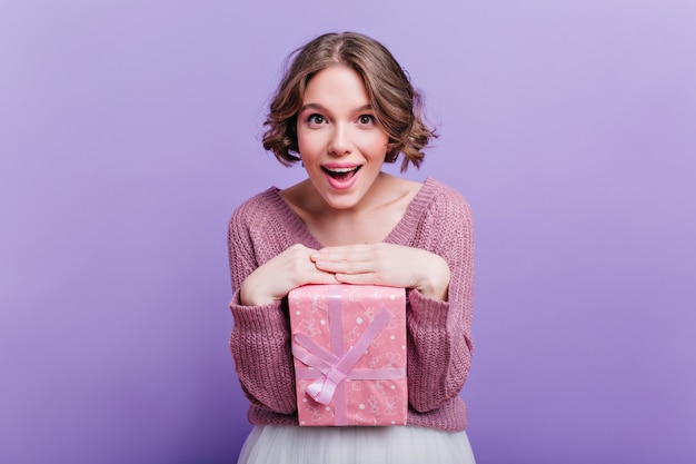Pleased short-haired woman posing with cute pink present box and smiling. enchanting curly girl enjoying photoshoot with new year gift on purple wall.