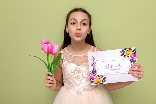 Pleased showing kiss gesture beautiful little girl on happy woman's day holding flowers with postcard isolated on olive green wall