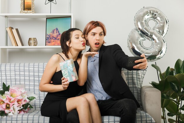 Pleased showing kiss gesture young couple on happy women day holding present take a selfie sitting on sofa in living room