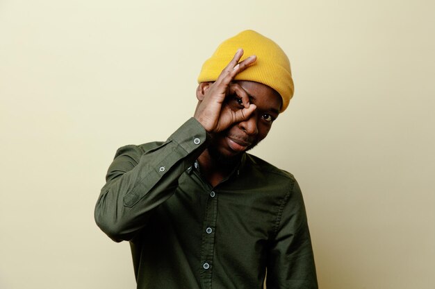 Pleased showing mask gesture young african american male in hat wearing green shirt isoloated on white background