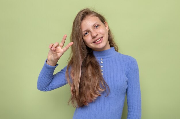 平和のジェスチャーを示すことを喜んでオリーブグリーンの壁に分離された青いセーターを着ている美しい少女