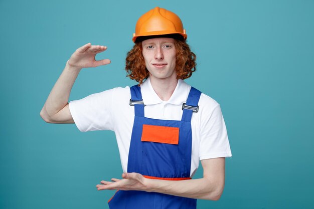 青い背景に分離された制服を着た若いビルダーの男性のサイズを示して喜んでいます