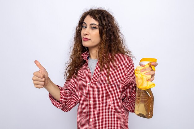 Pleased showing thumb up young cleaning woman holding cleaning agent with rag isolated on white wall