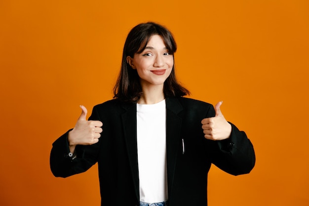 pleased showing thumbs up young beautiful female wearing black jacket isolated on orange background