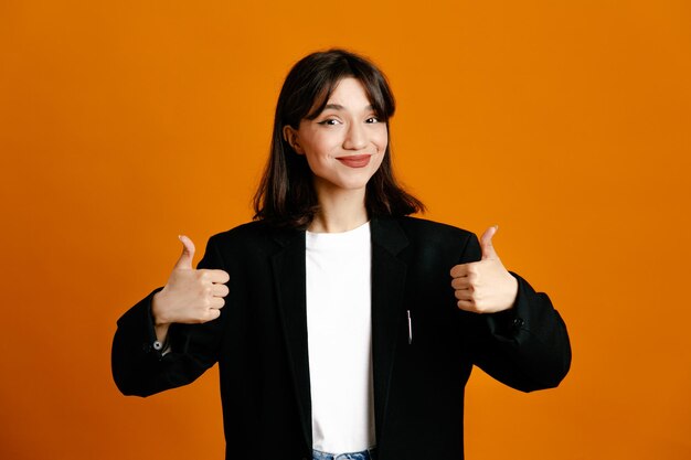 オレンジ色の背景に分離された黒いジャケットを着ている若い美しい女性を親指を示す満足