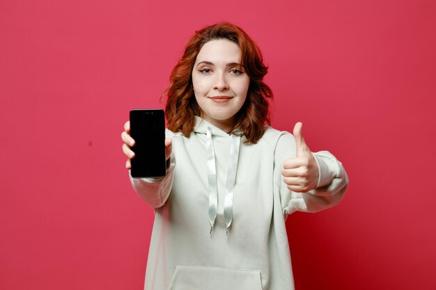 Pleased showing thumbs up young beautiful girl in white sweater holding phone isolated on pink background