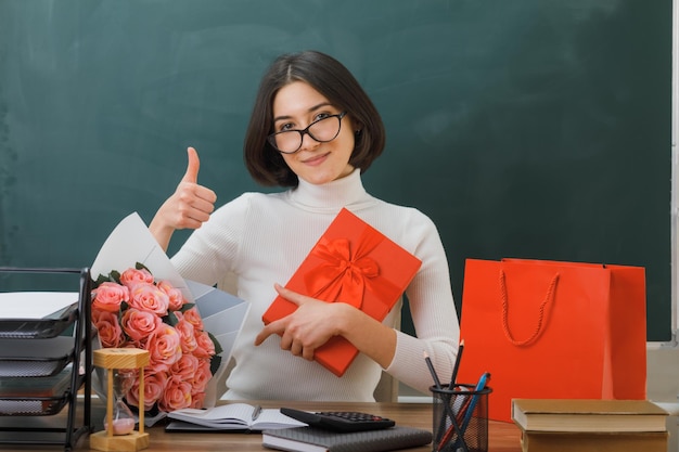 pleased showing thumbs up young female teacher holding present sitting at desk with school tools in classroom