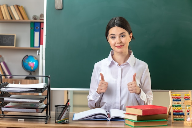 Lieto di mostrare i pollici in su giovane insegnante femminile seduta al tavolo con gli strumenti della scuola in classe