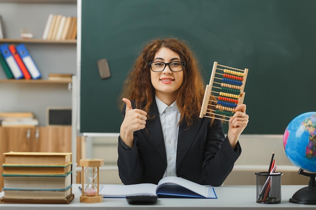 pleased showing thumbs up young female teacher wearing glasses holding abacus sitting at desk with school tools in classroom