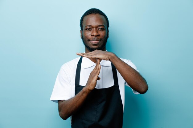 Pleased showing timeout gesture young african american barber in uniform isolated on blue background