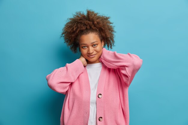 La donna timida e soddisfatta con i capelli afro tiene le mani sul collo e guarda direttamente soddisfatta. indossa un maglione rosa casual esprime emozioni sincere.