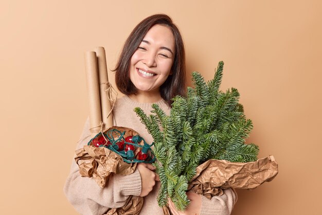 Pleased sincere Asian woman carries spruce branches rolled paper and retro garland tilts head smiles gently isolated over brown background People festive mood and holiday preparation concept