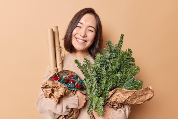 기쁘게 성실한 아시아 여성이 가문비나무 가지를 말아서 말린 종이와 복고풍 화환을 들고 갈색 배경 위에 부드럽게 고립된 머리 미소 사람들 축제 분위기와 휴가 준비 개념