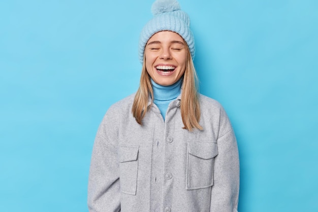 Pleased sincere young European woman keeps eyes closed smiles broadly expresses positive emotions wears knitted hat grey jacket isolated over blue studio wall. People clothes and fun concept