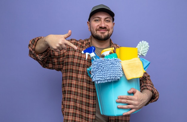 Pleased slavic cleaner man holding and pointing at cleaning equipment