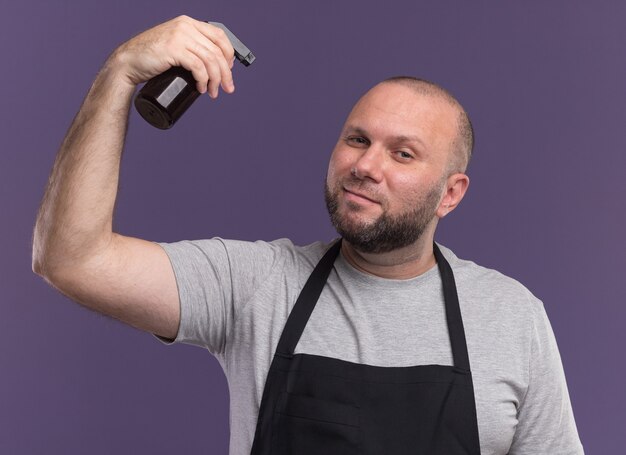 Pleased slavic middle-aged male barber in uniform raising spray bottle isolated on purple wall