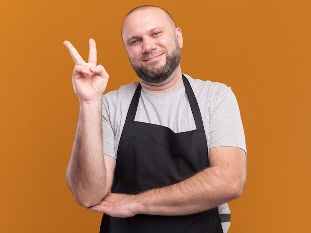 Free photo pleased slavic middle-aged male barber in uniform showing peace gesture isolated on orange wall