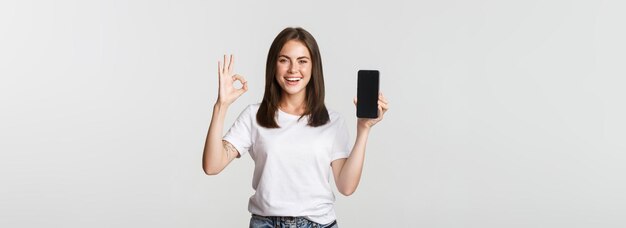 Free photo pleased smiling brunette girl showing smartphone screen and showing okay gesture in approval