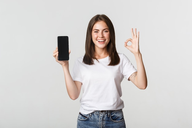 Pleased smiling brunette girl showing smartphone screen and showing okay gesture in approval.