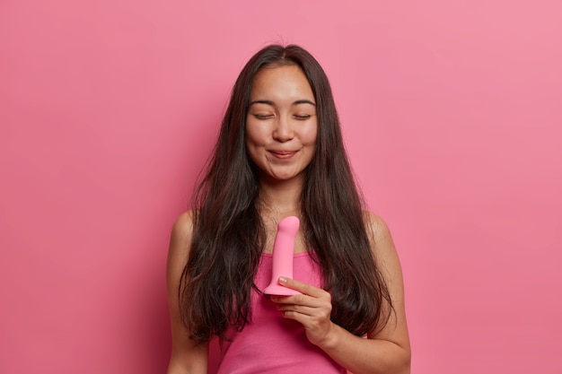 Pleased smiling brunette woman poses with silicone dildo for sexual penetration of vagina, mouth or anus, no need of partner, stimulates clitoris, uses sex toys for mastrubation, stands indoor