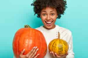 Free photo pleased smiling curly woman chooses pumpkin for halloween, holds big and small squash, wears white sweater