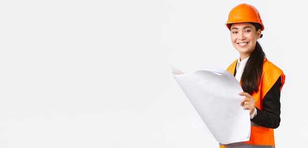 Pleased smiling female asian construction manager following blueprints during building house, looking satisfied at camera, studying architect plan, standing white background in safety helmet