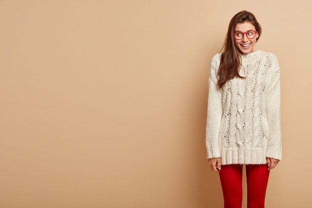 Free photo pleased smiling female wears spectacles, long white knitted jumper, red tights, looks aside, being in high spirit, stands over beige wall, blank space for your promotional content.