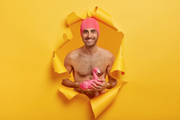 Pleased smiling man with naked torso, wears pink rubber swimcap, ready for summer vacation