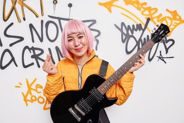 Pleased smiling pink haired teenage girl dreams to become rock star play music on acoustic guitar wears orange jakcet shows mini heart gesture or korean like sign poses against graffiti wall