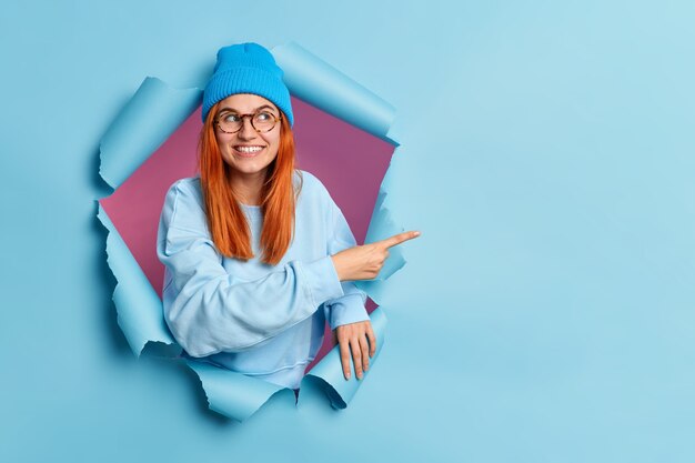 Pleased smiling redhead woman points finger at copy space shows special offer or shopping sale recommends good discount dressed in stylish blue outfit has happy mood breaks through paper hole