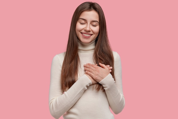 Pleased smiling woman keeps hands together on chest, expresses gratitude, keeps eyes closed