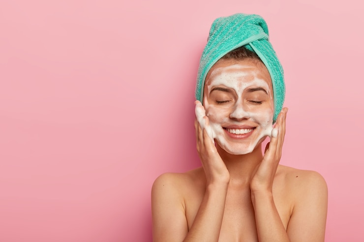 Free Photo pleased smiling woman washes face with cleansing gel, has soap on complexion, keeps eyes shut, wears wrapped towel on head, has naked body