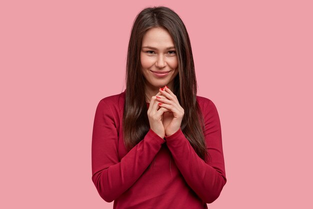 Pleased smiling woman with appealing appearance, keeps hands together near chin, wears red jumper, has intention to tell pleasant news to husband